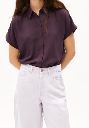 Persona con blusa de manga corta color púrpura oscuro, metida dentro de unos vaqueros gris claro de talle alto, de pie frente a un fondo blanco liso.