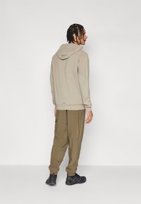 Bežs sporta krekls ar kapuci, brīvā piegriezumā, apvienots ar olīvu krāsas kravas biksēm un melniem sporta apaviem. Gluda tekstūra, vienkāršs dizains un elastīgas aproces.