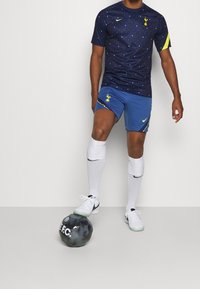 Maglietta sportiva blu navy con accenti gialli e motivo a pois, abbinata a pantaloni corti blu con logo. Calze bianche e palla nera.