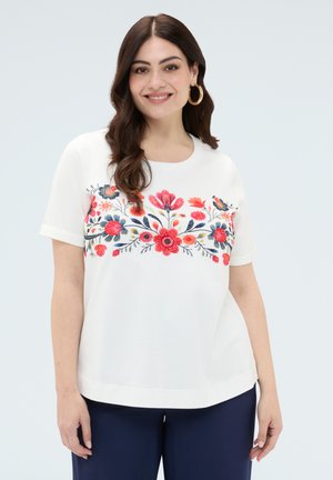 Kobieta z długimi brązowymi włosami nosi białą koszulę z czerwono-niebieskim haftem kwiatowym i granatowe spodnie, uśmiecha się do aparatu.