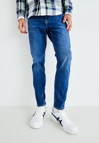 Vaqueros de mezclilla azul con un corte recto y un desgaste sutil, combinados con zapatillas blancas con acentos en azul marino y una suela texturizada.