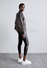 Brun läderbomberjacka med ribbade muddar, svarta figursydda byxor och vita sneakers mot en neutral bakgrund. Texturerade detaljer på jackan.