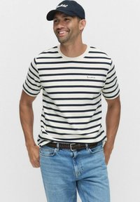 Hvid og marineblå stribet t-shirt med rund halsudskæring, korte ærmer og et lille logo på brystet, parret med lyseblå jeans og en mørk kasket.