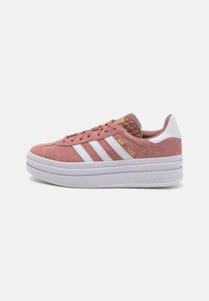 Baskets Adidas Gazelle en daim rose avec des bandes blanches et une semelle blanche épaisse sur un fond clair uni.
