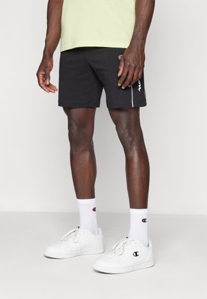 Man draagt zwarte Champion-shorts, witte Champion-sokken en witte Champion-sneakers en staat op een effen achtergrond.
