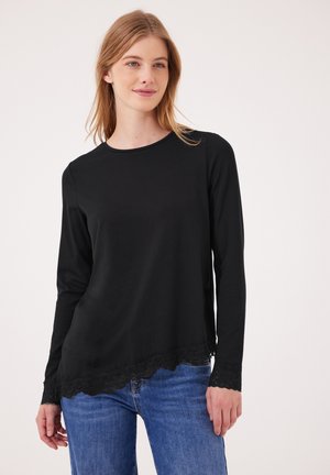 Jeune femme portant un haut noir à manches longues avec bordure en dentelle et un jean bleu, debout devant un fond clair uni.