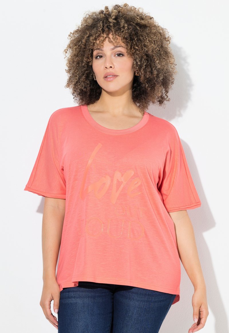 Mujer con cabello rizado, vestida con una camiseta suelta color coral que dice "Love out Loud" y jeans de mezclilla azul oscuro, de pie contra un fondo blanco.