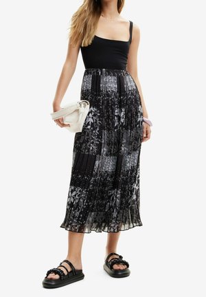 Vrouw draagt een zwarte mouwloze top en een geplooide zwart-witte midi-rok met bloemmotief, houdt een witte handtas vast en draagt zwarte sandalen.