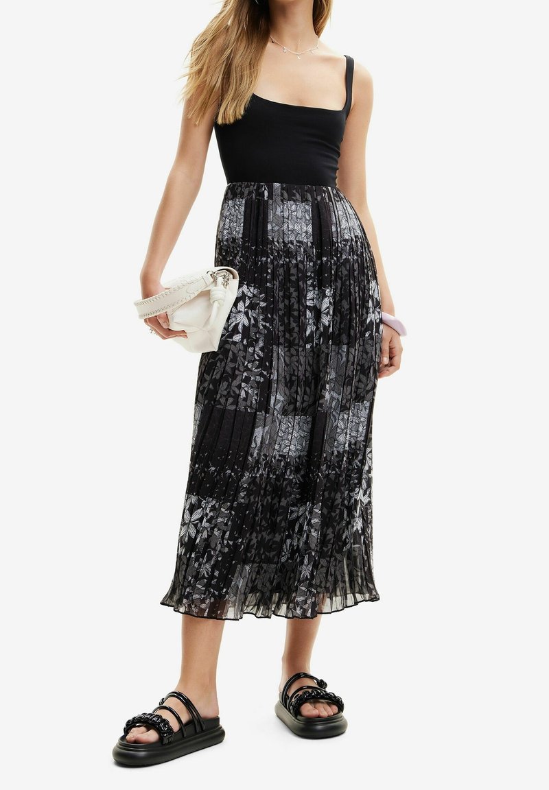 Femme portant un haut noir sans manches et une jupe midi plissée à motifs floraux noirs et blancs, tenant un sac à main blanc et portant des sandales noires.