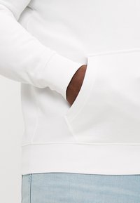 Biela mikina s dlhými rukávmi, priliehavými manžetami a klokankovým vreckom, zobrazená proti svetlému džínsovému materiálu. Mäkka textúra látky.