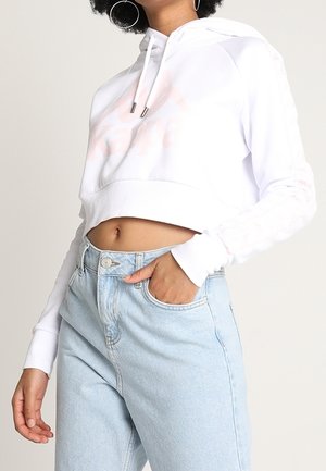 Frau trägt einen kurzen weißen Hoodie mit Kordeln und hellblauen High-Waist-Jeans, Hand in der Tasche, neutraler Hintergrund.