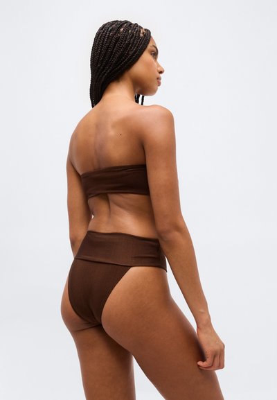 Kobieta z plecionymi włosami, ubrana w brązowy top bandeau bez ramiączek i bikini z wysokim stanem, stojąca bokiem na jednolitym tle.