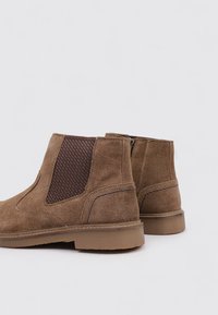 Braune Wildleder-Stiefeletten mit elastischen Seitenpaneelen, strukturierten Stoffakzenten und einer niedrigen Gummisohle. Merkmale sind Stitching-Details im Obermaterial.