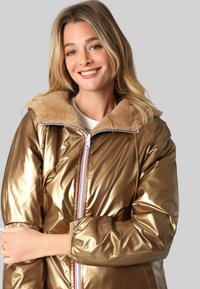 Veste rembourrée en métal doré avec un col en fausse fourrure, fermeture éclair à l'avant et accents côtelés. Présente une finition brillante et une silhouette décontractée.