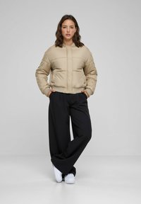 Beige puffjacka utan dragkedja framtill och med ribbat nederkant, i kombination med vida svarta byxor och vita sneakers, placerad mot en neutral bakgrund.