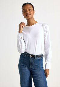 Maglietta bianca a maniche lunghe con scollatura rotonda, maniche arricciate e dettagli con bottoni, abbinata a jeans blu a vita alta e una cintura nera.