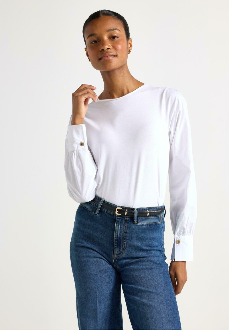 Maglietta bianca a maniche lunghe con scollatura rotonda, maniche arricciate e dettagli con bottoni, abbinata a jeans blu a vita alta e una cintura nera.