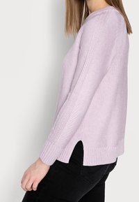 Suéter de punto lila claro con un acabado texturizado, aberturas laterales y puños de canalé, combinado con jeans negros.