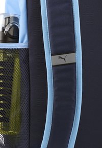 Zaino blu navy con accenti azzurri, dotato di una tasca in rete che contiene una borraccia nera. Logo Puma su una patch grigia. Materiali testurizzati.
