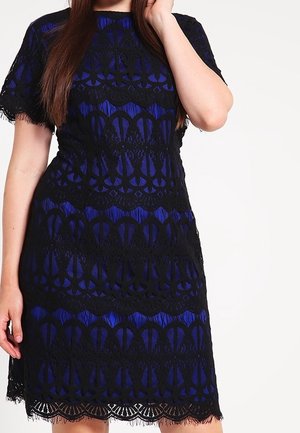 Woman wearing a knee-length royal blue dress with black lace overlay, short sleeves, and scalloped hem, standing against a white background.
