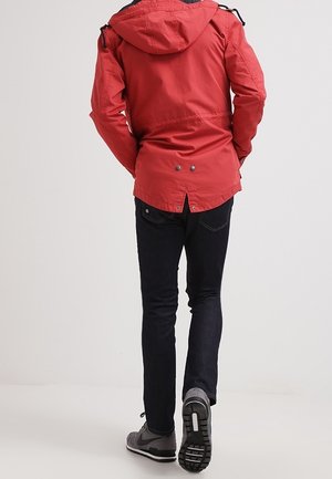 Personne portant une veste à capuche rouge, un jean foncé et des baskets grises marchant sur un fond clair uni.