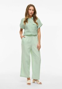 Completo a due pezzi in tessuto verde chiaro; camicia a maniche corte con bottoni e tasca, pantaloni a gamba larga e sandali con tacco di colore chiaro.