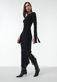 Schwarzes, geripptes Strickkleid mit langen, ausgestellten Ärmeln, verziert mit goldenen Akzenten und Schlitzen am Saum. Getragen mit schwarzen, spitz zulaufenden Stiefeln.