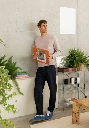Man in casual kleding die een oranje vinylplaathoes vasthoudt, staand in een moderne kamer met planten, boeken en een platenspeler.
