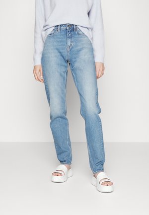 Persoon draagt lichtblauwe high-waisted spijkerbroek, witte trui en witte platform sandalen, staand tegen een effen witte achtergrond.