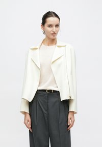 Hvid uldblandet jakke med reverskrave og asymmetrisk lynlås, båret over en lyserød top og mørkegrå højtaljede bukser.