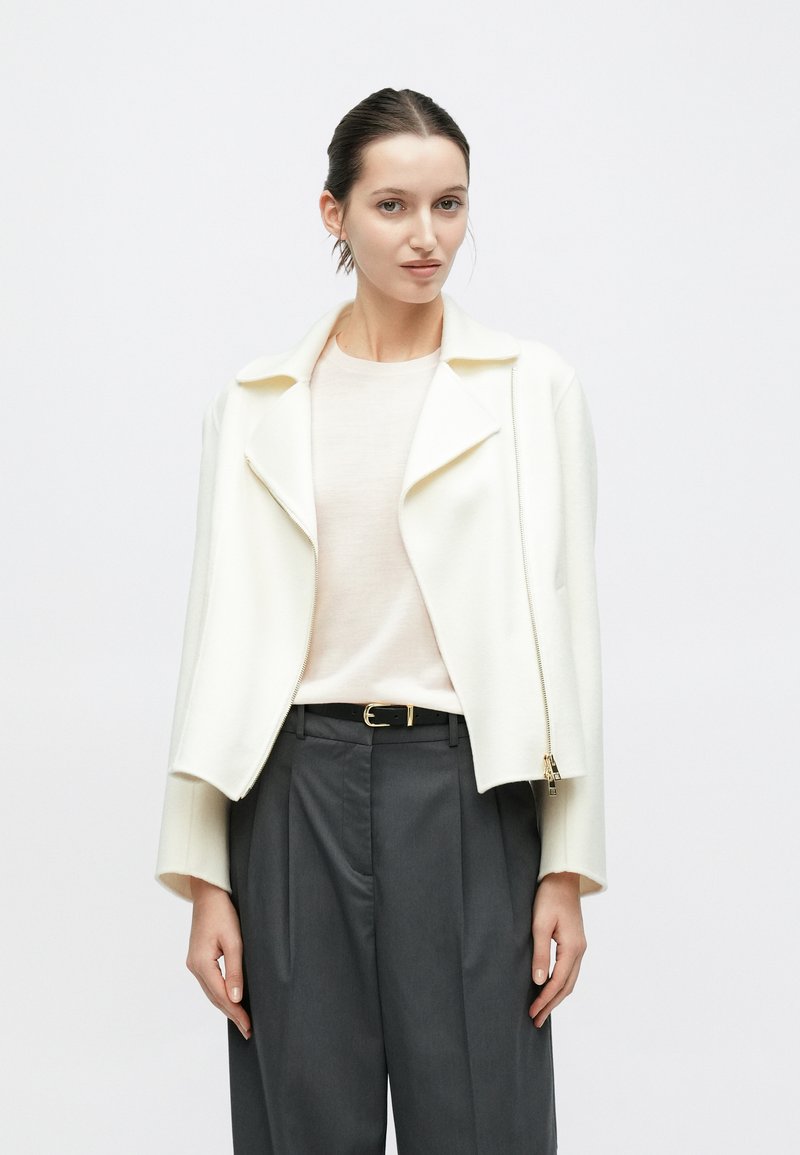 Giacca in misto lana bianca con colletto a revers e cerniera asimmetrica, indossata sopra una maglietta rosa chiaro e pantaloni grigio scuro a vita alta.