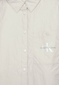 Lichtgrijs overhemd met een gestructureerde afwerking, voorzien van een klein borstzakje en een logo erop gedrukt. De knopen zijn gegraveerd met 'CK'.