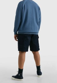 Blaues Textursweatshirt mit gerippten Bündchen, kombiniert mit dunklen Shorts mit Seitentaschen. Modell trägt dunkle Sneakers und Knöchelsocken.