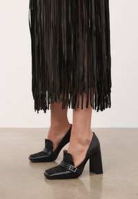 Fekete bőr magassarkú cipők texturált elülső panellel és csat részletekkel, hosszú fekete fröccsenő szoknyával viselve egy betonpadlón.