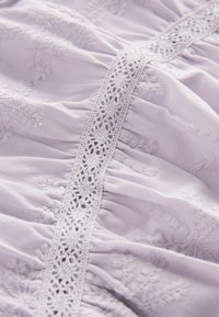 Tissu lavande avec broderie texturée et délicate dentelle, présentant un ruché pour plus de profondeur et de dimension.