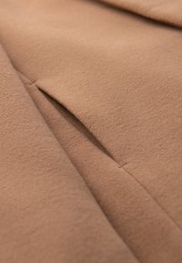 Close-up af et beige uldstof, der viser en pænt syet søm og en åbning til en slidslomme.