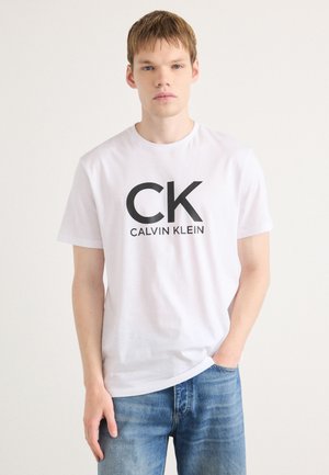 White cotton t-shirt featuring a bold black "CK" logo and "CALVIN KLEIN" text underneath, paired with blue denim jeans.