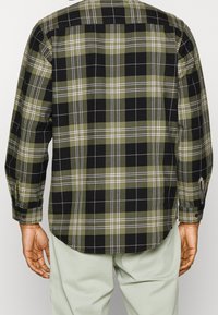 Chemise à carreaux verts et noirs avec manches longues, poignets boutonnés et ourlet droit. Fabriquée en tissu texturé avec un motif à carreaux.