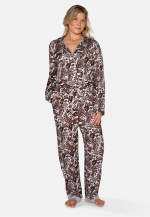 Lyshåret kvinde stående barfodet iført løstsiddende pyjamas med paisleyprint, knap-op top og bukser med snøre på en ensfarvet hvid baggrund.