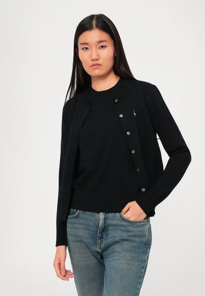 Una donna con lunghi capelli scuri indossa un cardigan nero con bottoni sopra un top nero e jeans blu, in piedi su uno sfondo semplice chiaro.