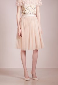 Gonna di tulle beige con vita arricciata, abbinata a un corpetto ricamato con fiori. L'outfit presenta un top trasparente a strati e tacchi nudi.