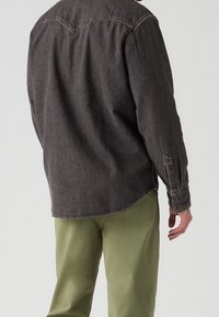 Persona che indossa una camicia di jeans a maniche lunghe grigio scuro e pantaloni verde oliva chiaro, vista da dietro su uno sfondo semplice.
