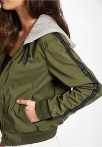 Olivegrön bomberjacka med grå fleecehuva, svart logotape på ärmarna, framfickor och ribbade muddar och fåll.