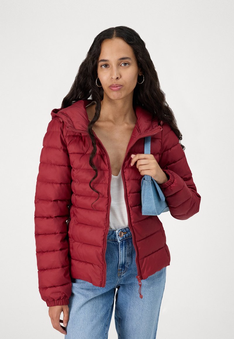 Veste matelassée rouge avec un quilting horizontal, fermeture éclair et capuche réglable. Associée à un haut léger et un jean bleu taille haute.