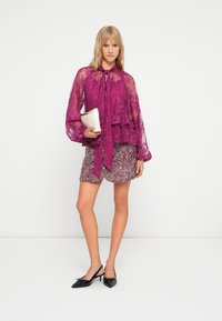 Fuksia bluse i blonder med spennehals og ballongsleeves, kombinert med et mønstret miniskjørt. Holder en hvit clutch og har på seg svarte leiligheter.