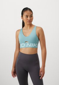 Svijetloplava sportska grudnjak s prorezom sprijeda, s širokim pojasom na kojem je ispis "PRO NIKE". Materijal izgleda glatko, nosi se s visokim sivi tajicama.