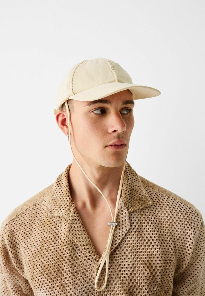 Beige Kappe mit einem gebogenen Schirm, gestickten Nähten und verstellbarem Riemen. Kombiniert mit einem hellbraunen, strukturierten Strickhemd.
