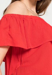 Top rosso scollato sulle spalle con collo arricciato, realizzato in tessuto leggero, caratterizzato da una superficie strutturata e un bordo elastico.