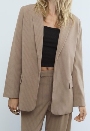 Beige pinstripe bleiseri rento leikkauksella, yksirivinen malli ja lovet kaulukset. Puettu mustan topin päälle, yhdistettynä samanvärisiin housuihin.