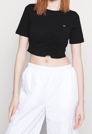 Femme portant un crop top noir froncé ajusté et un pantalon blanc ample avec une ceinture élastique, debout devant un fond clair uni.
